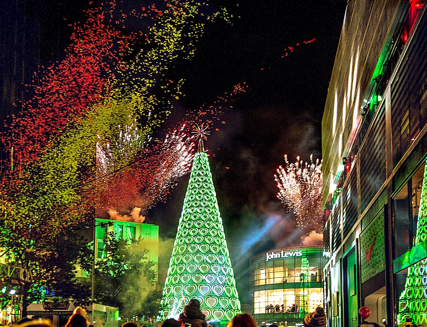 Celebratory fireworks display above a decorated Christmas tree in an urban setting at night, with a crowd of people watching.