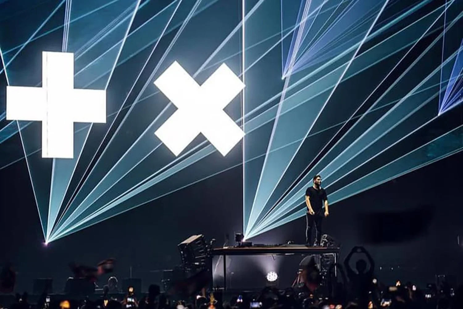 A DJ performs on stage with a giant screen behind him. The screen displays three symbols: a plus sign, an 'X', and a circle, all in white. Light beams and lasers project outward from the stage, illuminating the dark background, with an audience in front taking photos.