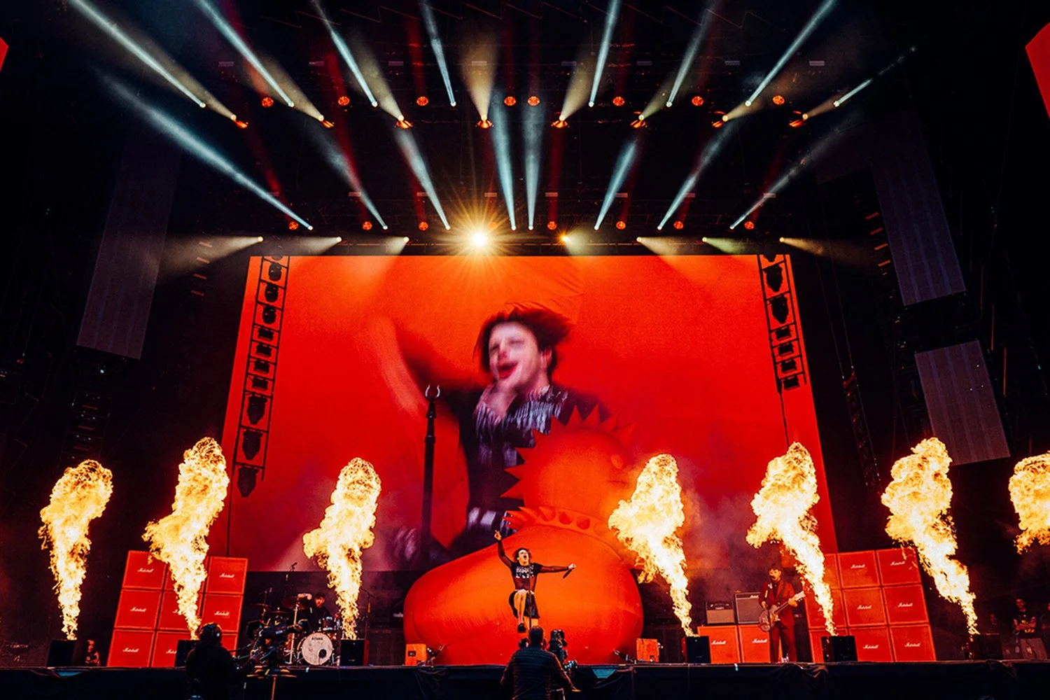 A concert stage with bright spotlights, large video screen, flames, and musicians performing.