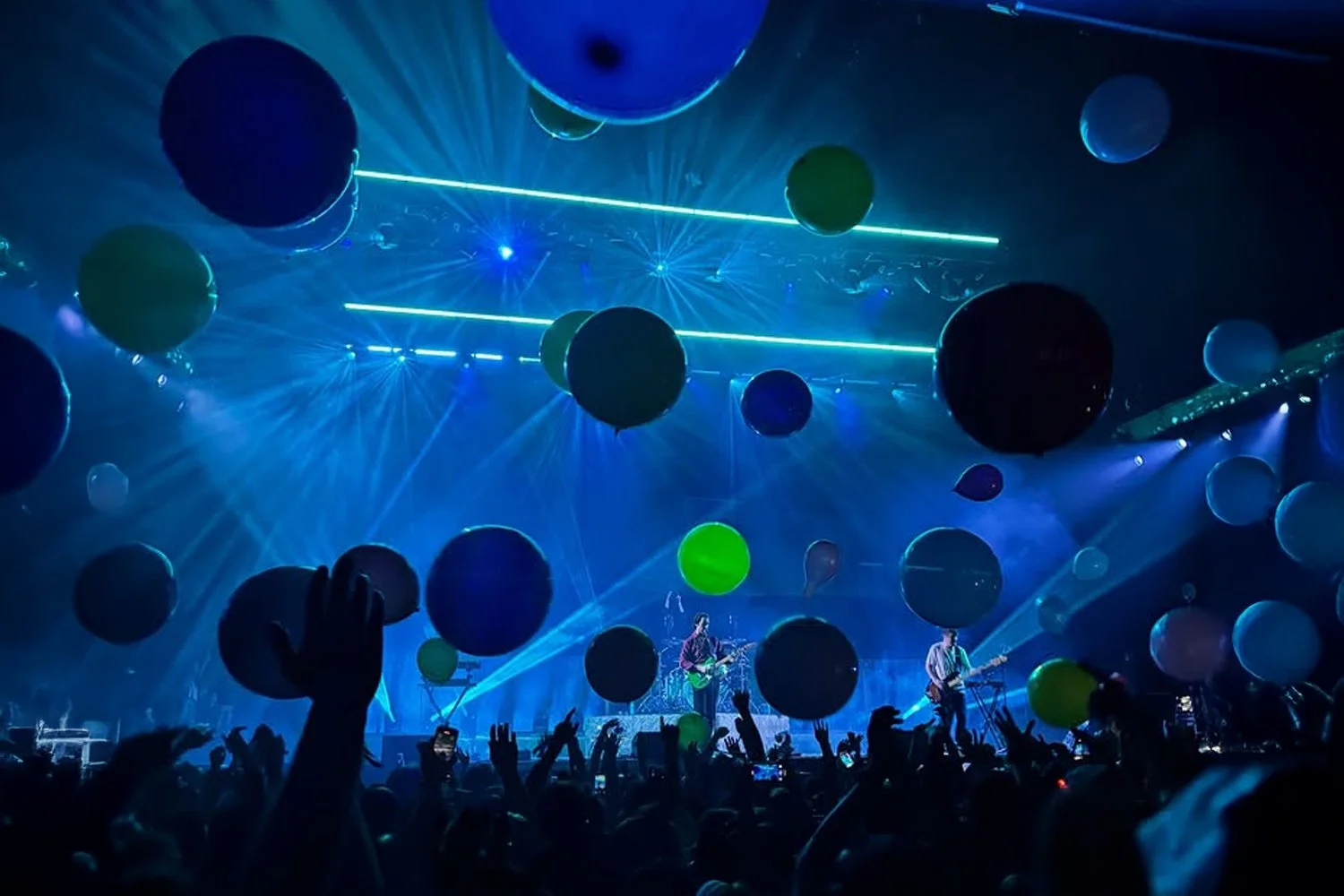 A concert scene with a band playing on stage, surrounded by large balloons in green, blue, black, and white, suspended in the air. Audience members are seen raising their hands, some holding phones, with colorful laser lights and blue lighting creating a vibrant atmosphere.