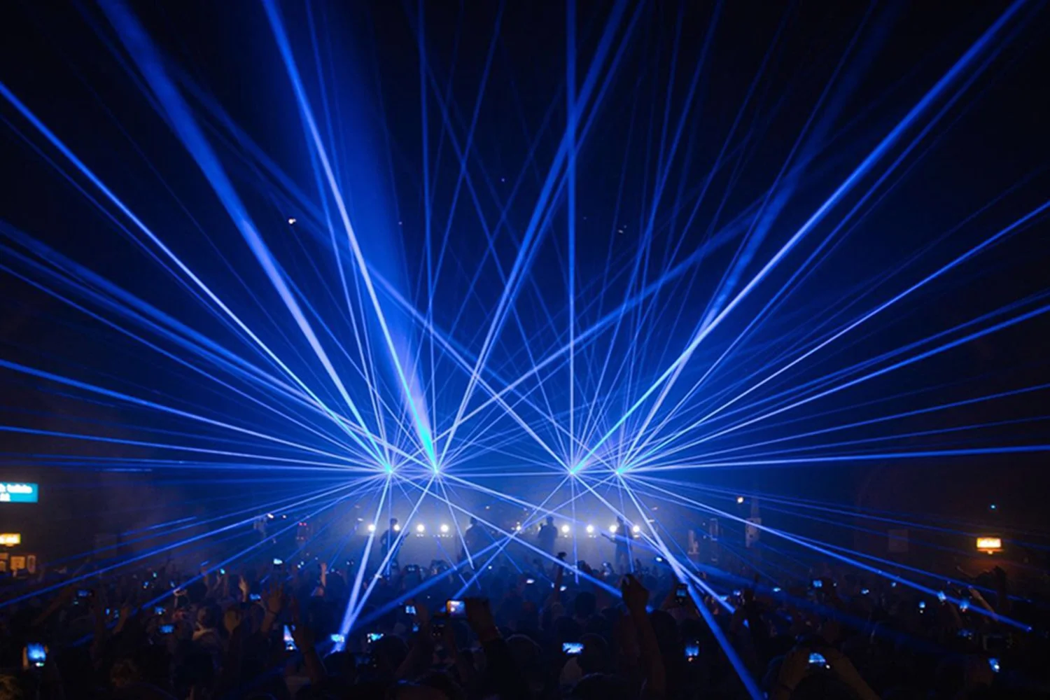 Blue laser beams projected over a crowd at a concert or event, with silhouettes of people taking pictures.