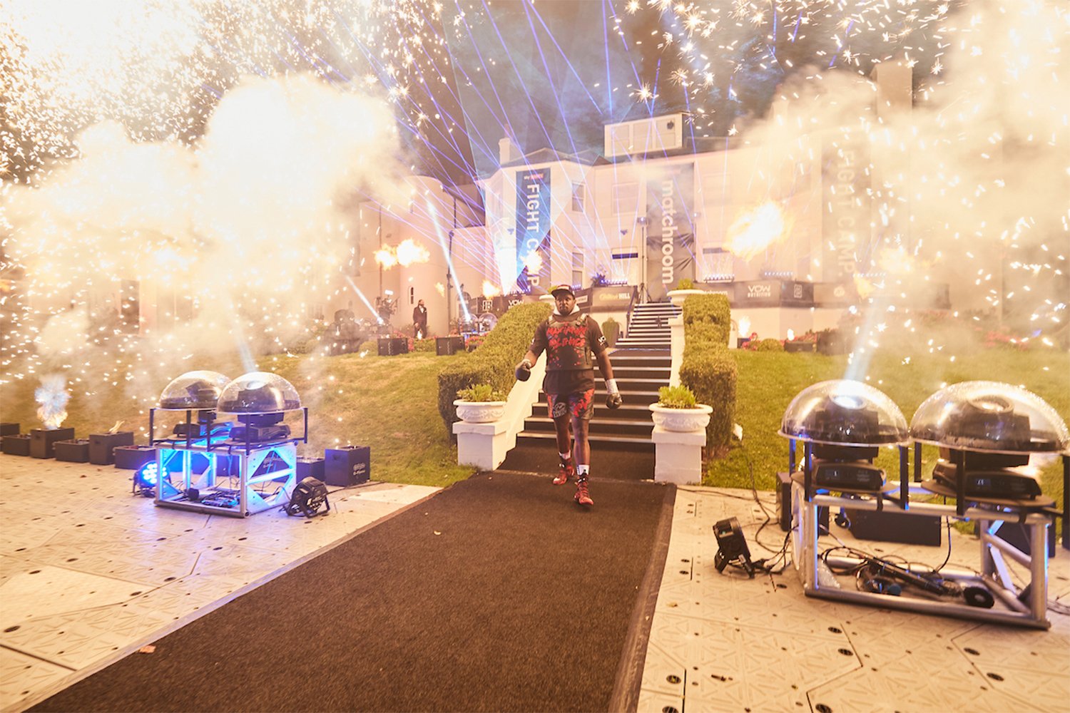 A fighter walks through a fireworks display outside a modern building with illuminated signs and banners, during a celebration or event at night.