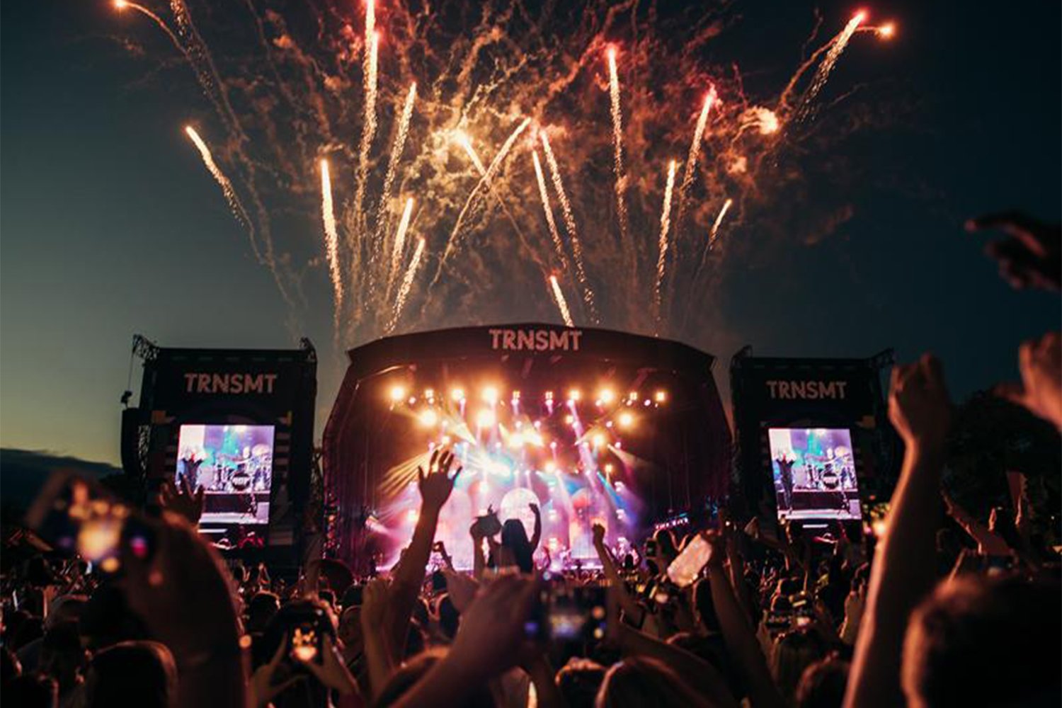 Concert at night with fireworks overhead, large crowd raising hands, stage with bright lights, and screens showing performers, at an outdoor music festival called TRNSMT.