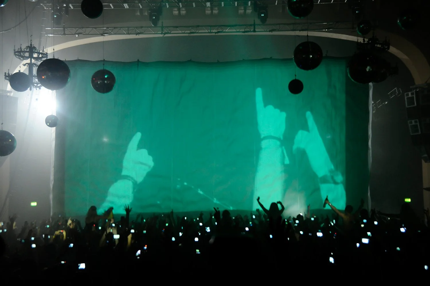 Silhouettes of people at a concert or event, with a large screen showing an image of hands making gestures, and hanging spherical decorations in the venue.