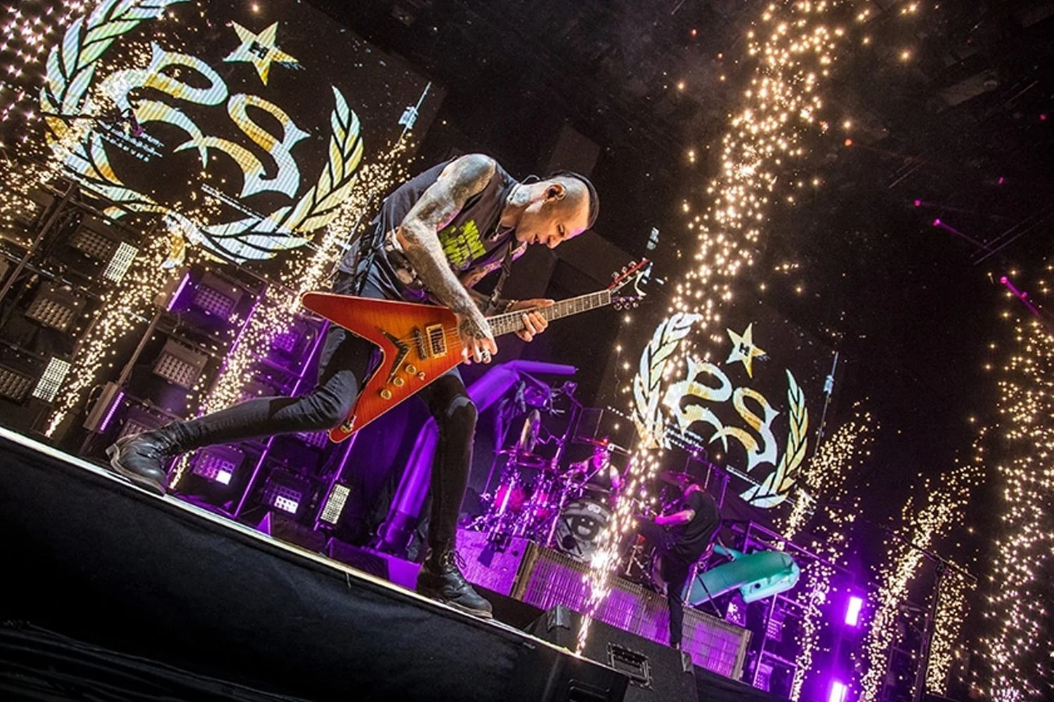 A guitarist performing on stage during a concert with pyrotechnics and fireworks, with a large screen behind displaying a logo with a star and laurel wreath.