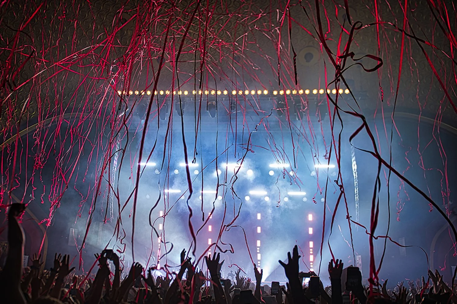Concert crowd with hands raised, confetti falling, stage with bright lights and smoke.