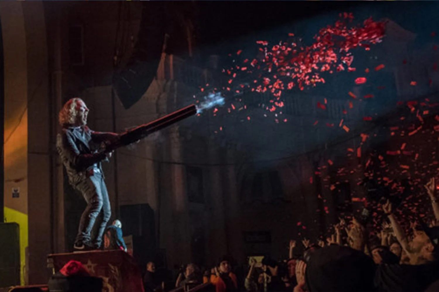 Person standing on a platform, holding a confetti cannon, releasing pink confetti into the air. Crowds of people below watching the celebration.