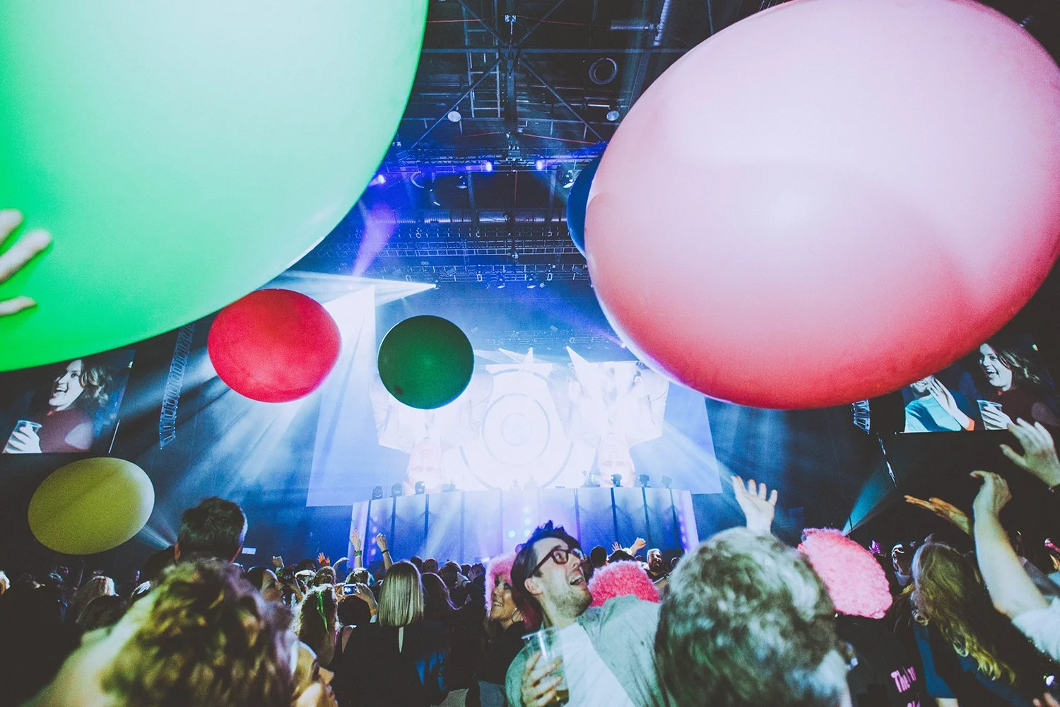 People at a concert or event with large colorful balloons hanging overhead, stage with lights and a screen in the background, and attendees enjoying and dancing.