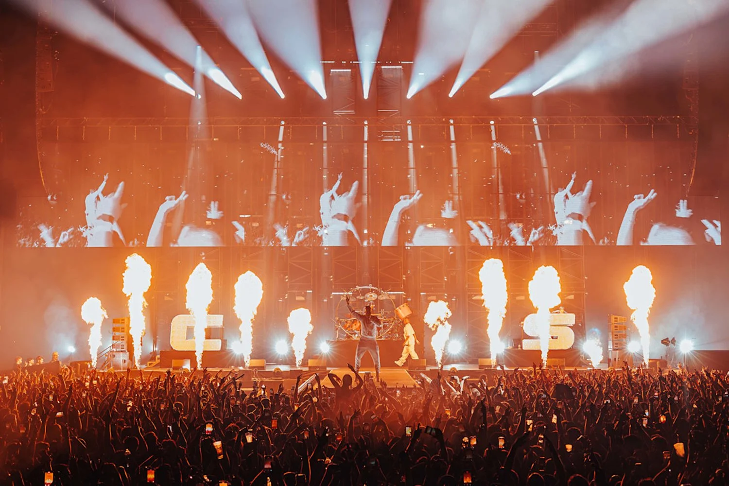 Concert stage with performers, pyrotechnics, large video screen showing hands and a DJ, and an audience with many people holding phones