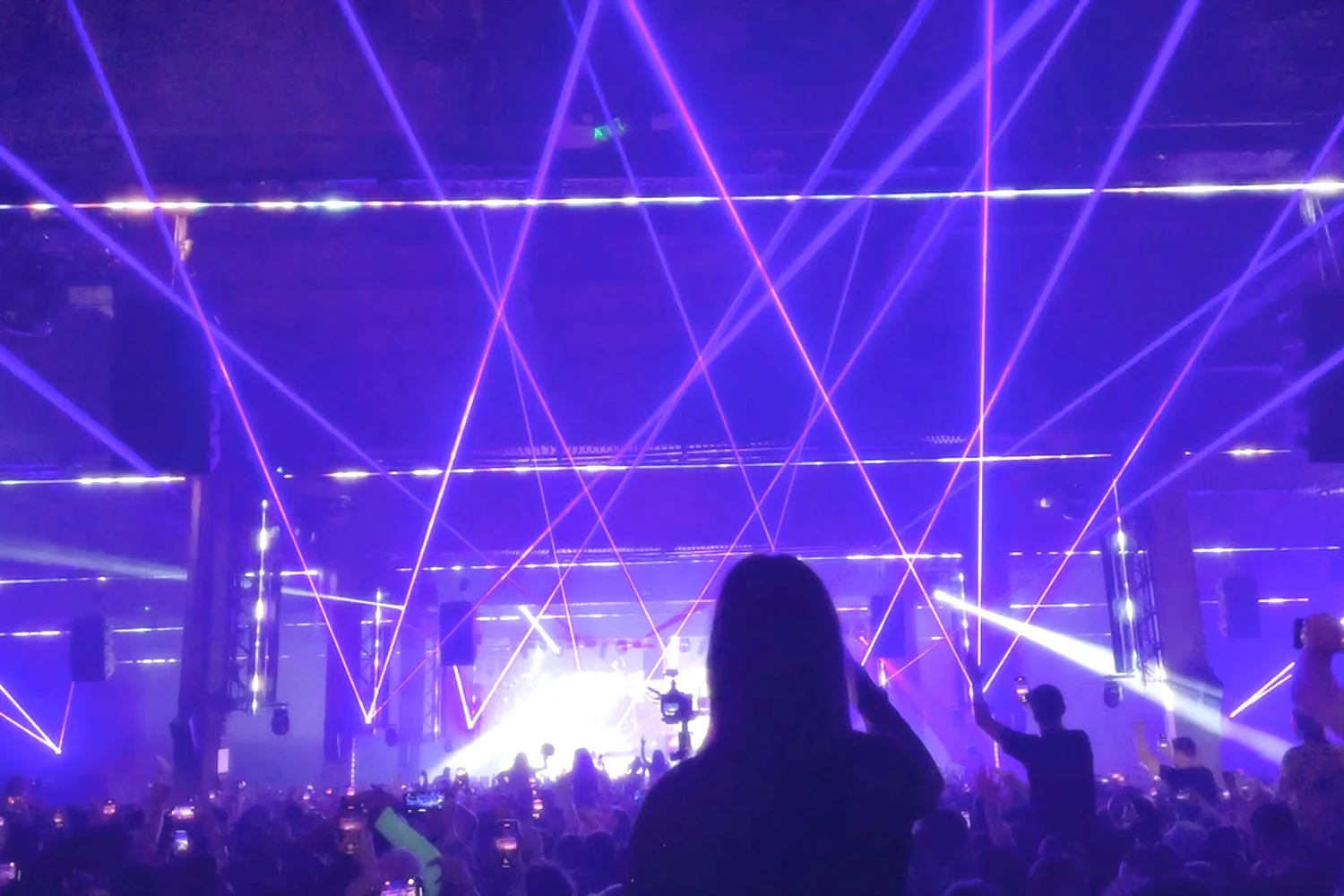 Crowd at a concert with purple laser lights crossing the stage and audience taking photos or videos.