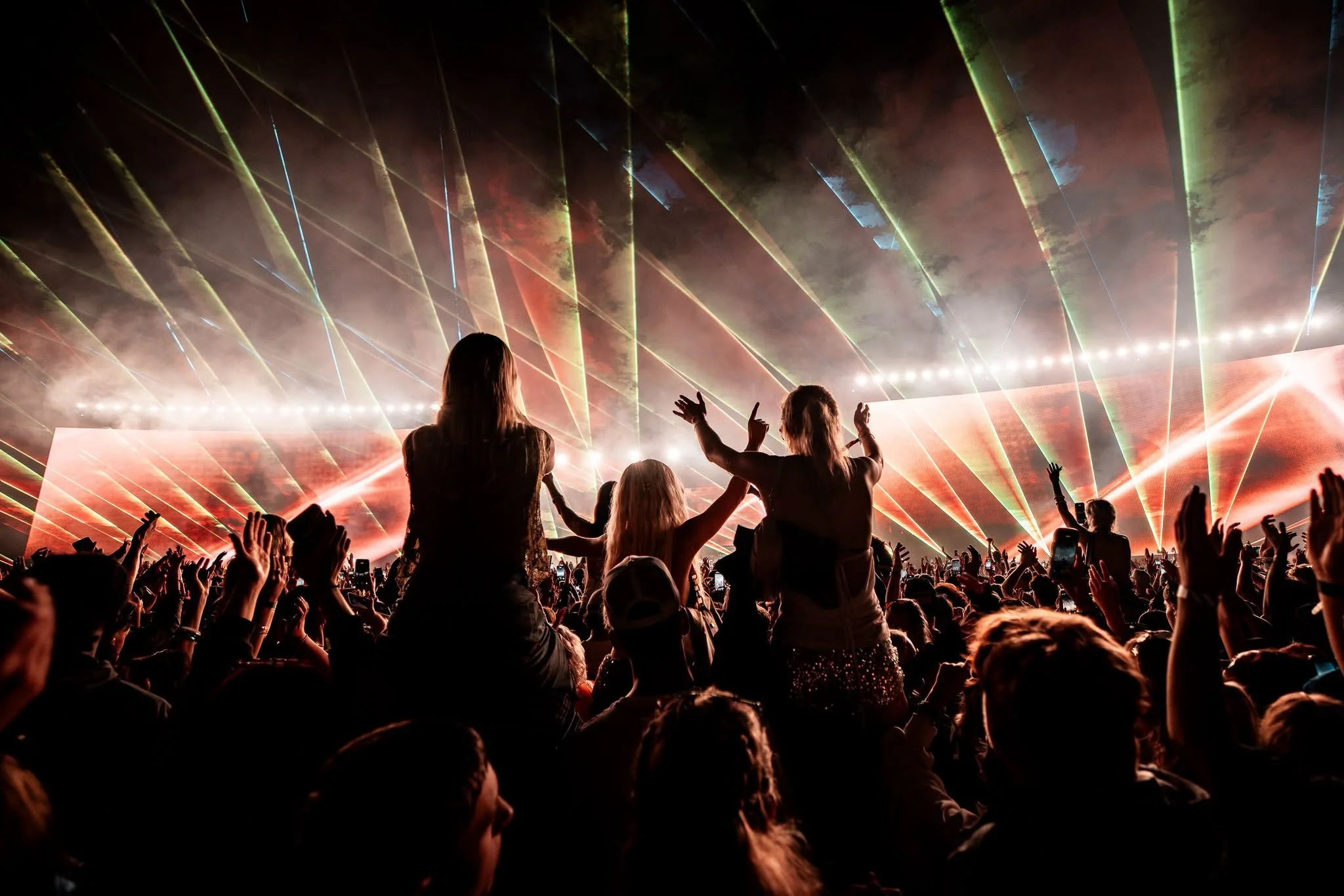 A large crowd at a concert with people dancing and raising their hands, with colorful laser lights and a foggy atmosphere.