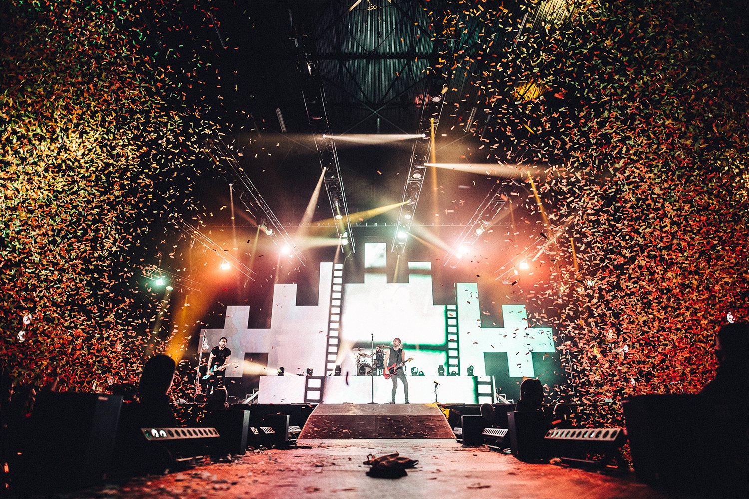Concert stage with band performing, colorful lighting, yellow and white spotlights, digital screens, and confetti falling.