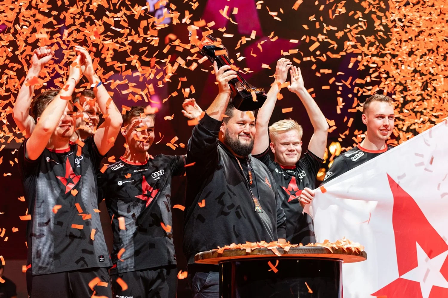 Esports team celebrating victory with confetti, some team members clapping, one holding a trophy, and another holding a flag, all wearing matching team jerseys.
