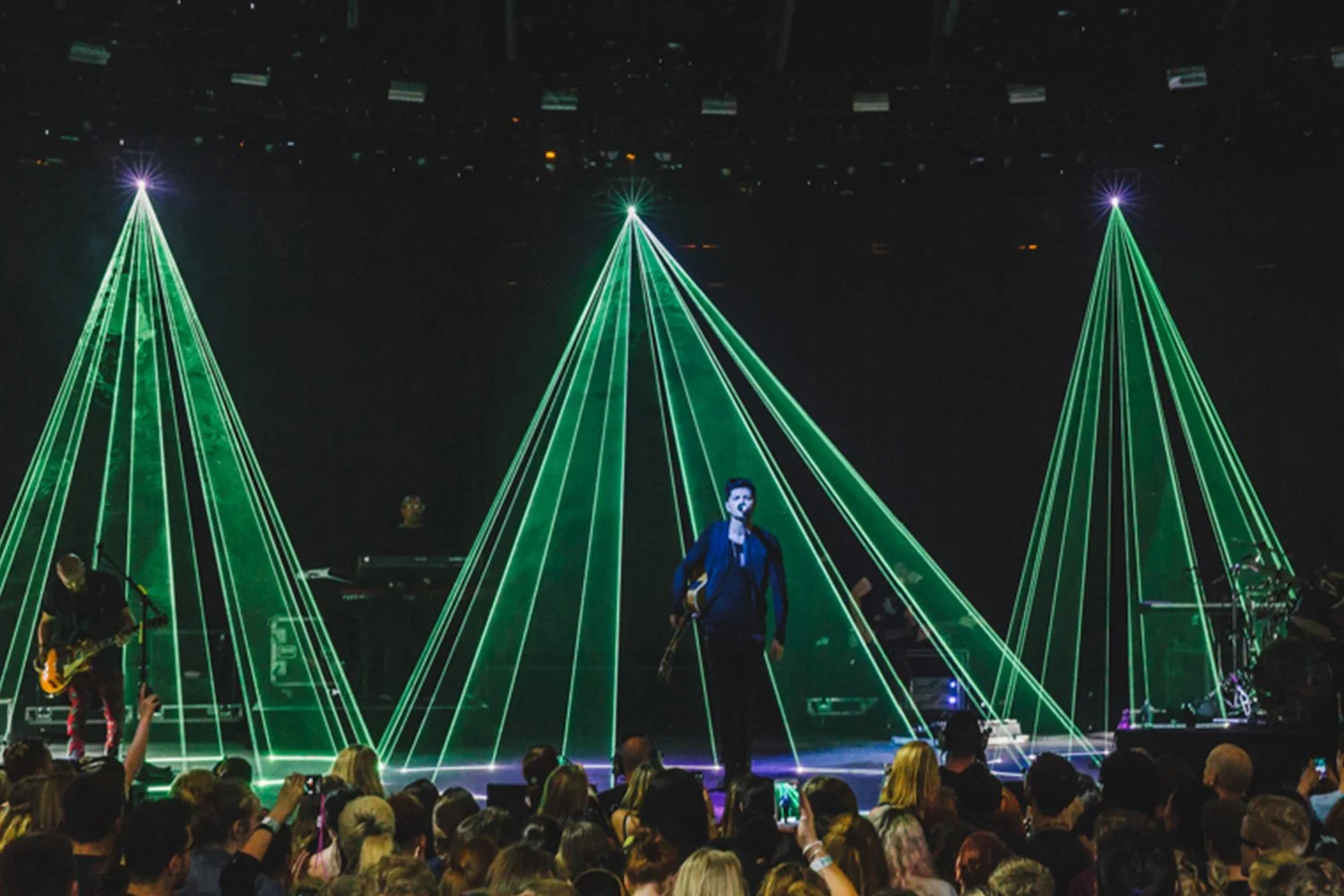 Musician on stage with green laser light display forming triangular patterns, band members playing instruments, audience watching and taking photos.
