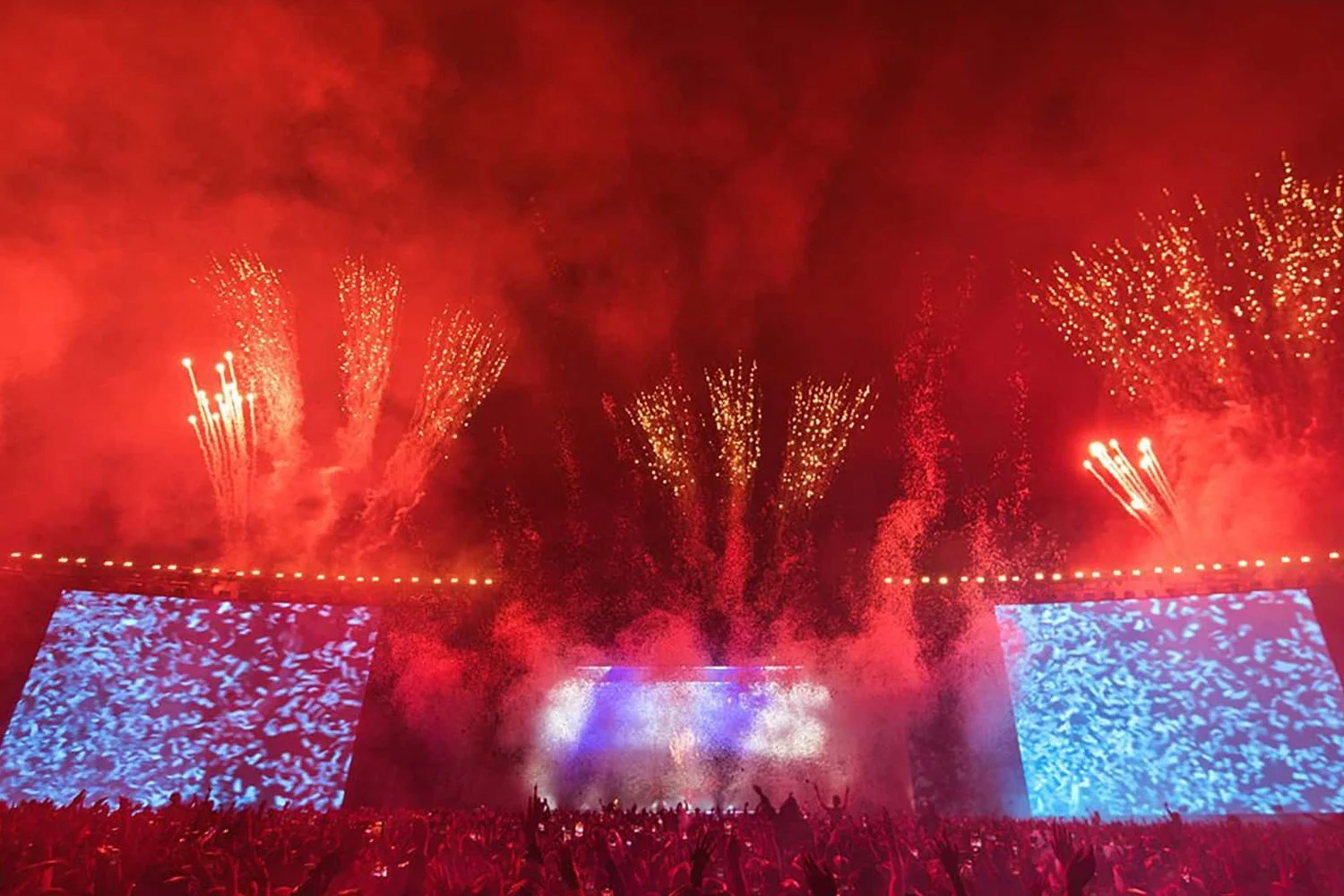 Large outdoor concert with fireworks display, red sky, and bright screens showing blue patterns, audience with raised hands.