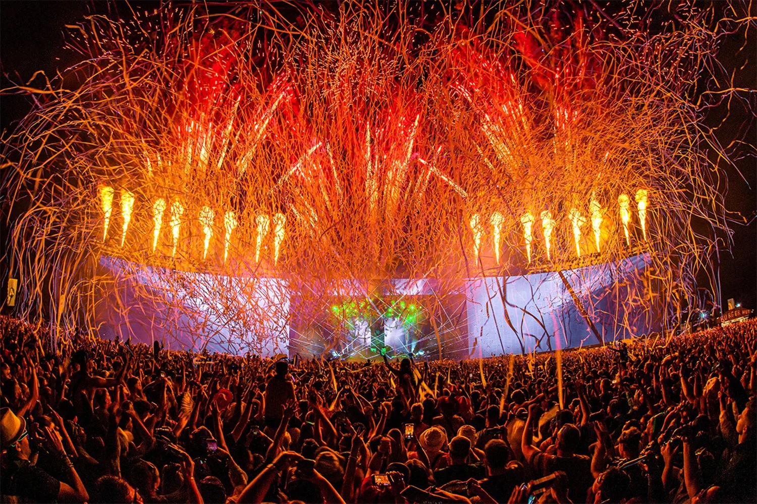 A large crowd at a concert or festival during night with fireworks and confetti in the sky and stage lights.