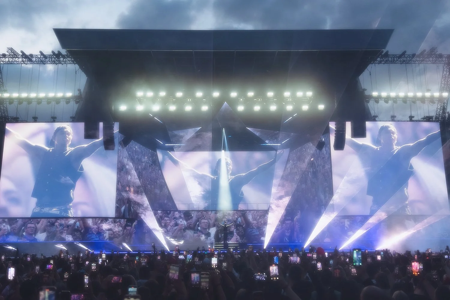 A large outdoor concert stage with a performer in front, screens displaying the performer, and an audience with many people taking photos with their phones. The sky is cloudy.