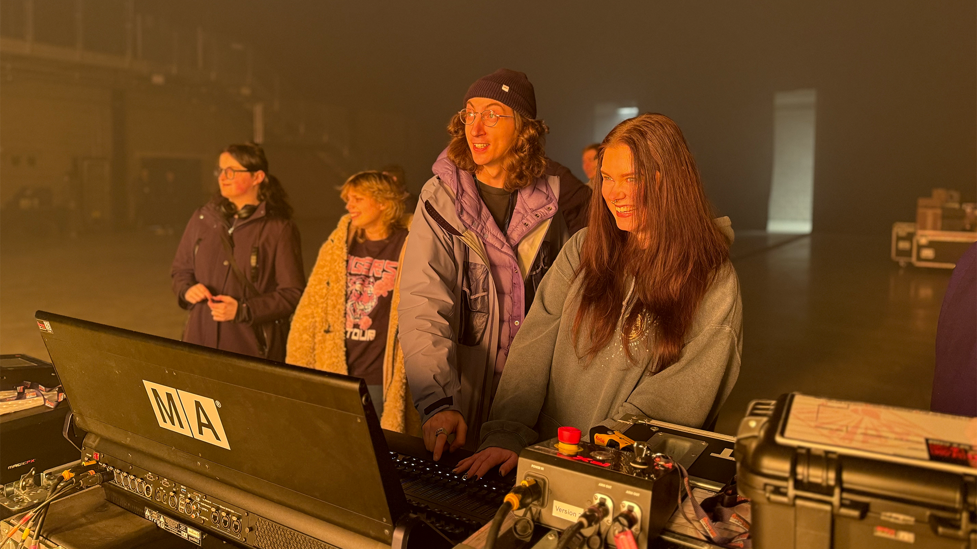 Group of people on a film set in a large indoor space with equipment. The group appears to be preparing or rehearsing, with a focus on two women smiling and interacting near a control panel.
