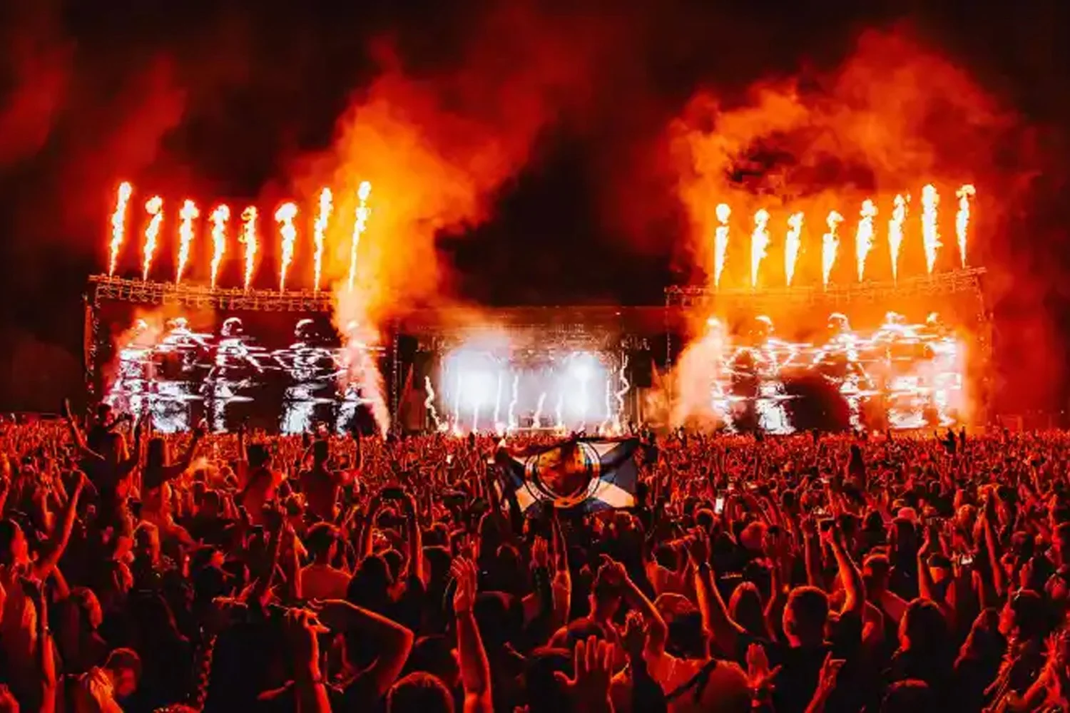Crowd at a concert with a large stage, bright lights, and flames shooting up from the stage, creating a fiery display during the night.