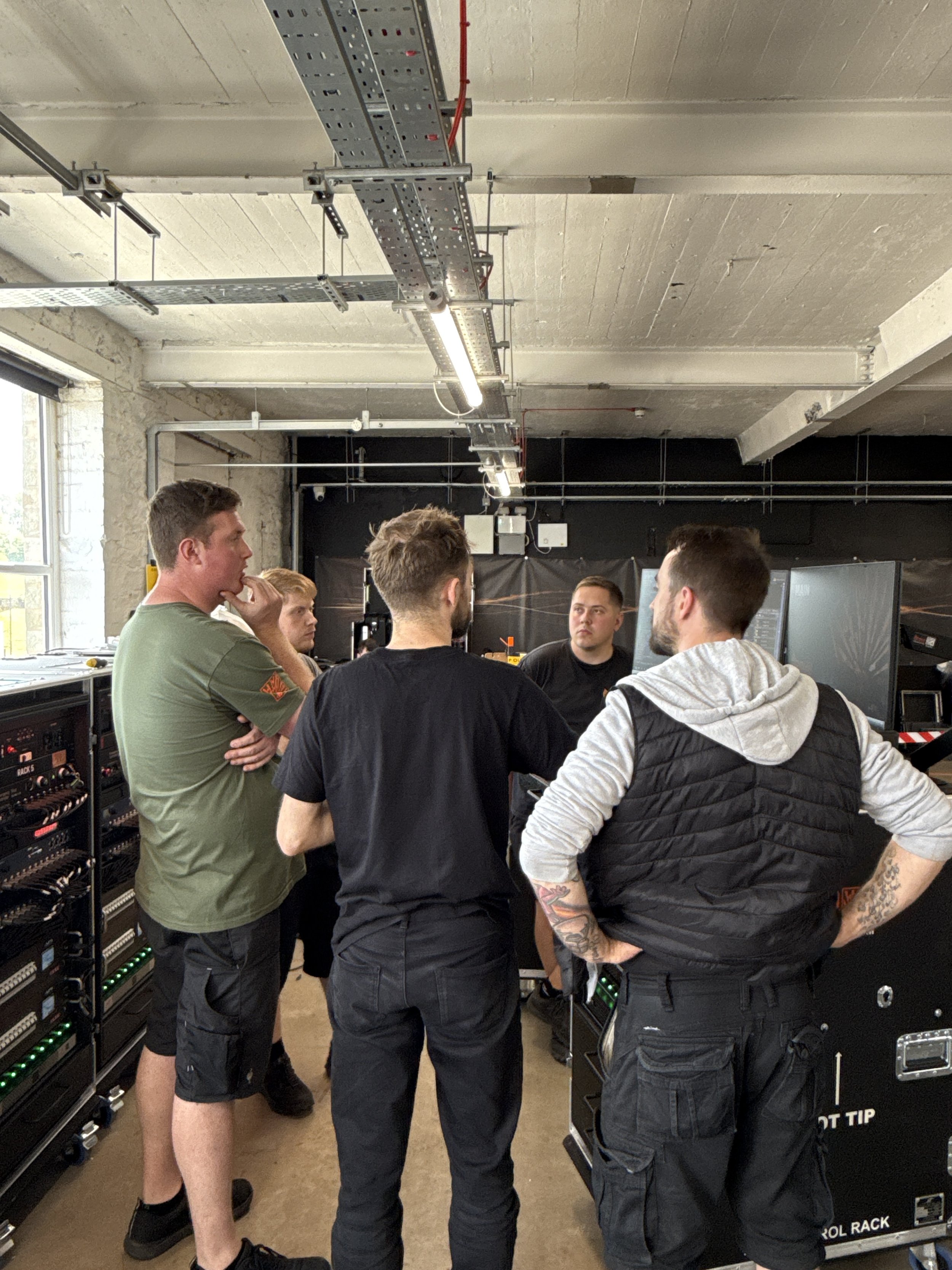 Group of young men standing in a server room with computer equipment and cabling on racks.