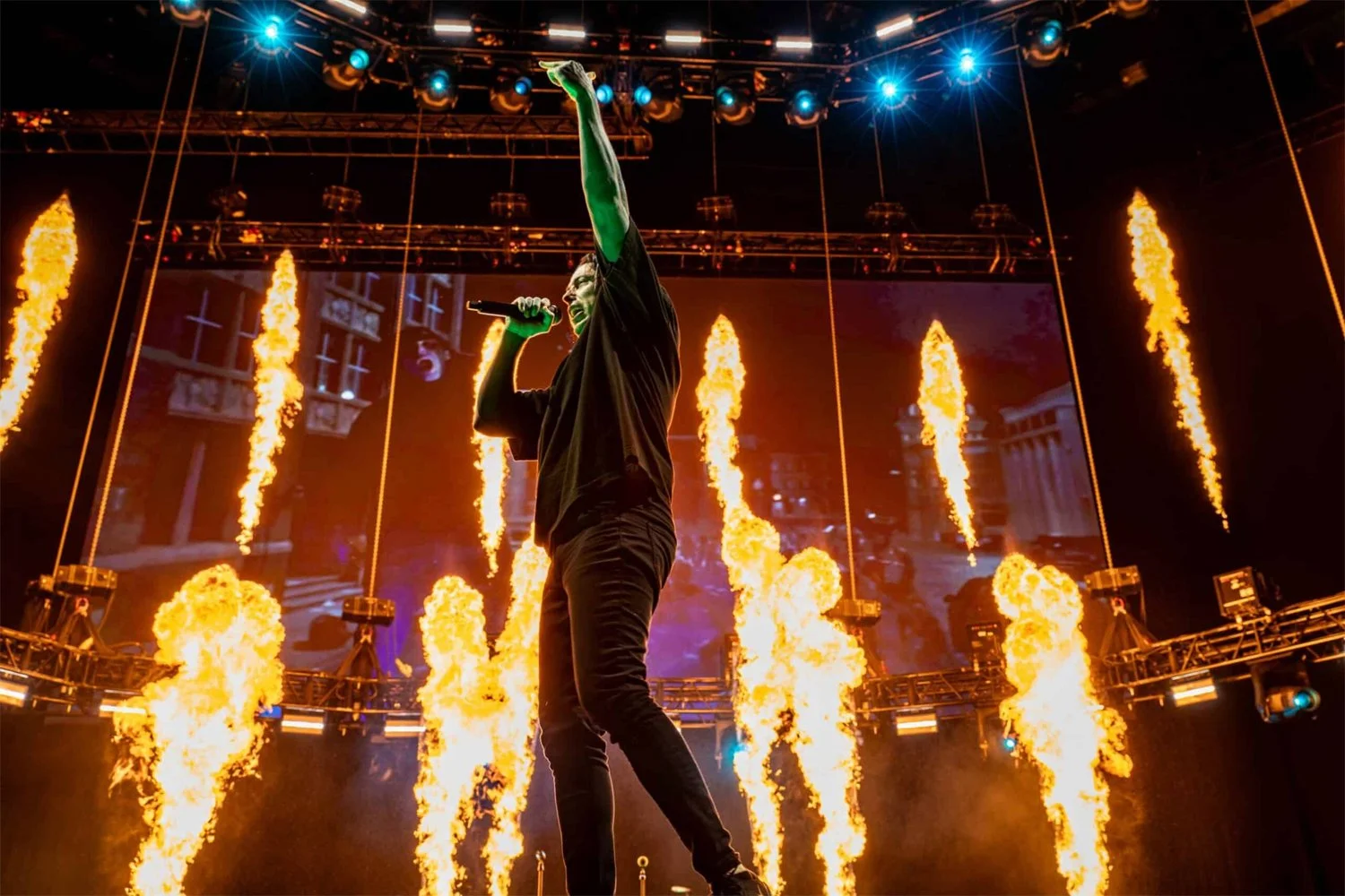 Performer singing on stage with flames shooting upward behind him and blue lights overhead.