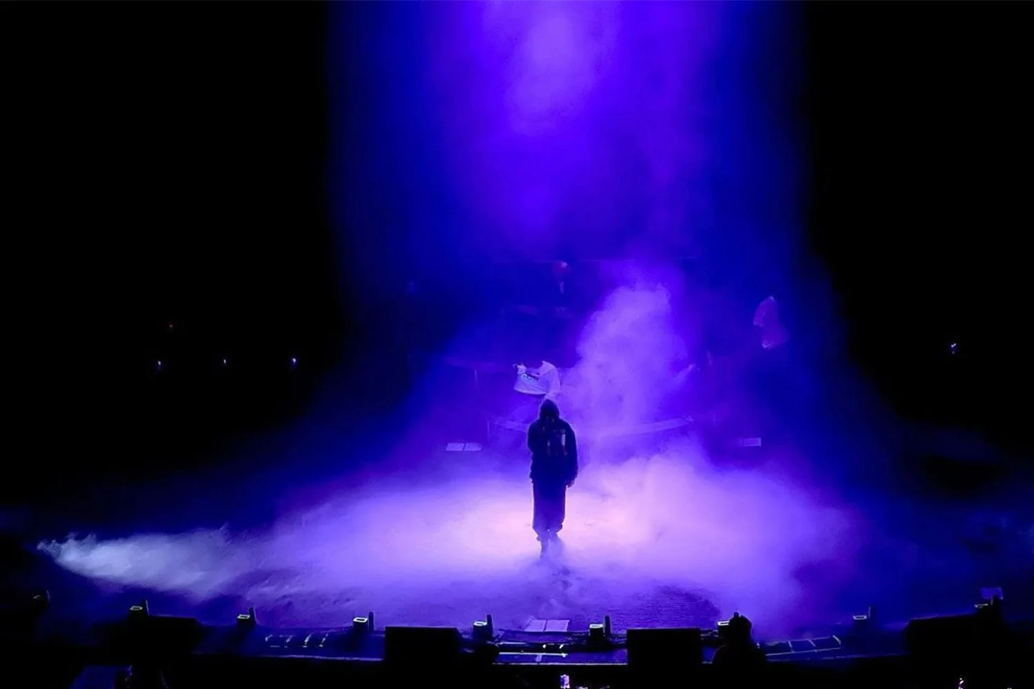 A person standing in the center of a foggy stage illuminated by purple light, with a flag in hand, surrounded by mist and darkness.