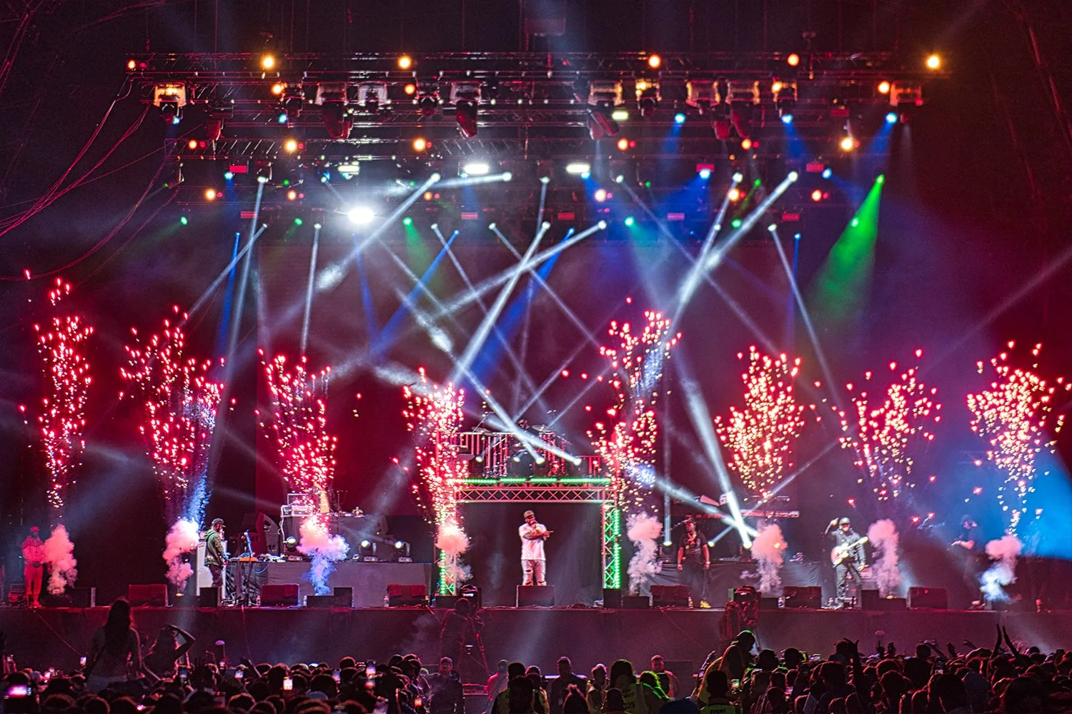 Concert stage with performers, colorful lights, fireworks, and an audience.
