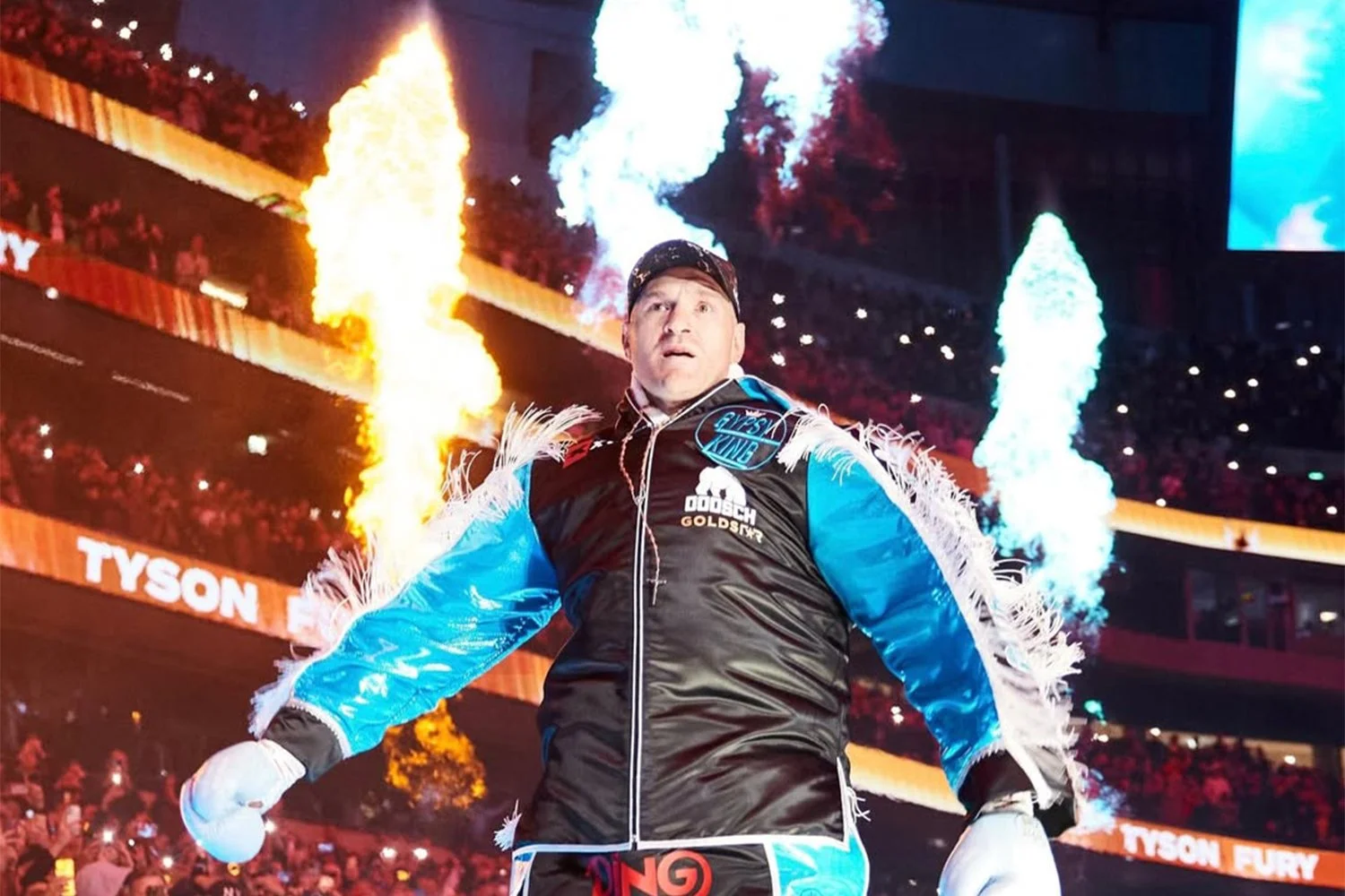 A boxer stands in the ring with flames and fire effects behind him, wearing a black, blue, and white outfit, with the words Tyson Fury visible on the background signs.