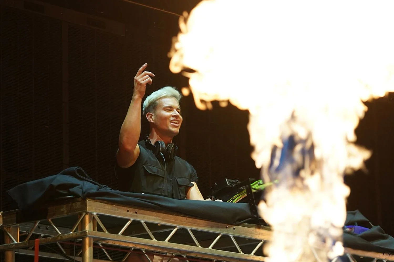 A DJ with platinum blonde hair smiling and raising his hand while standing behind a DJ booth with fire in the foreground.
