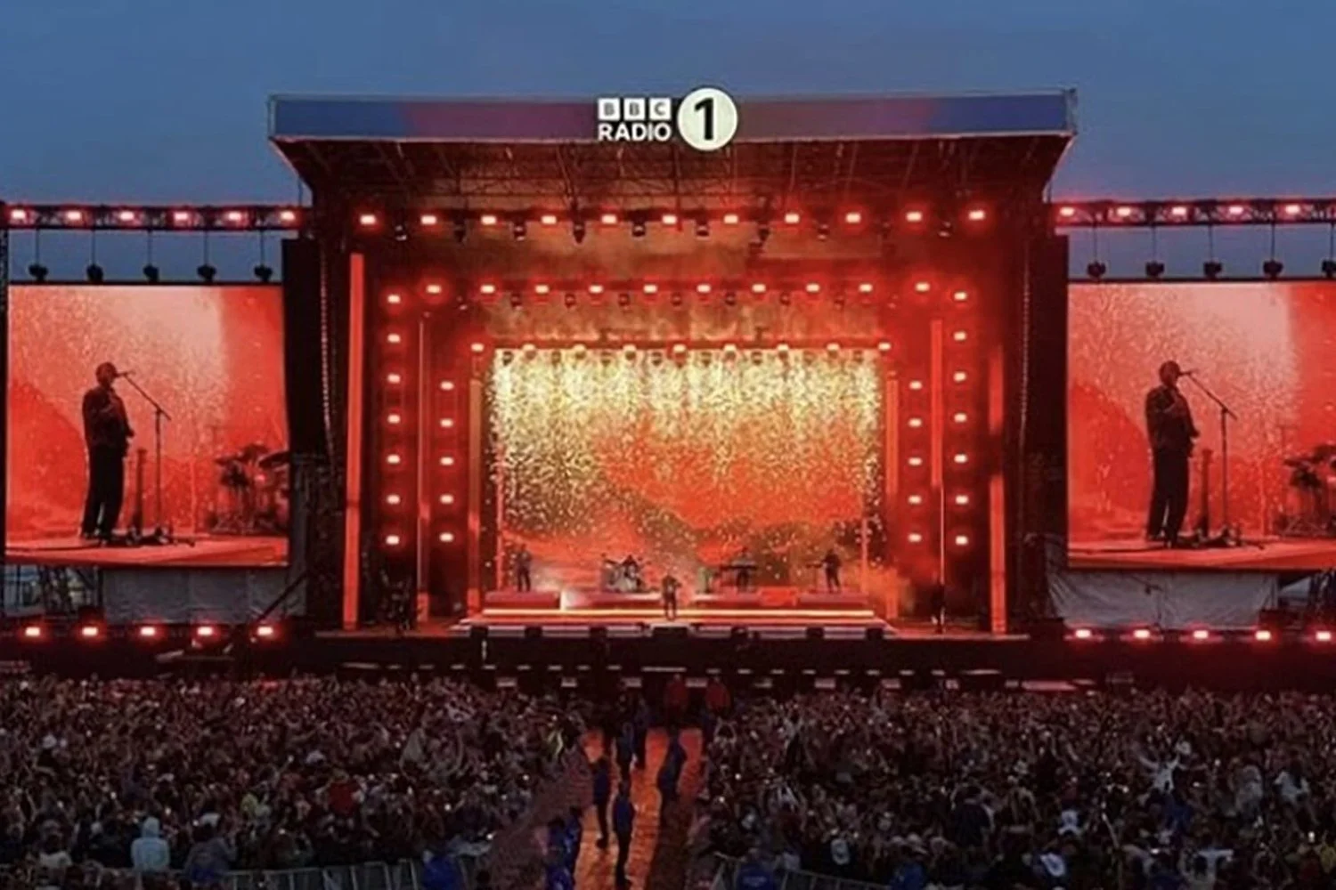 A large outdoor concert stage with red and orange lighting, featuring a band performing with two large screens on either side and an audience watching in the evening
