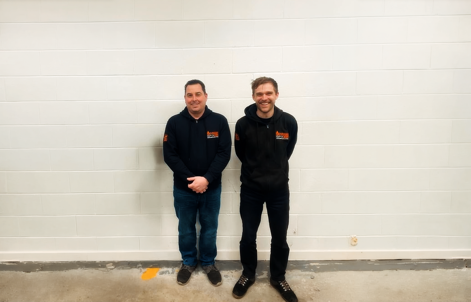 Two men standing against a white wall, both wearing black zip-up hoodies with orange and white logos.