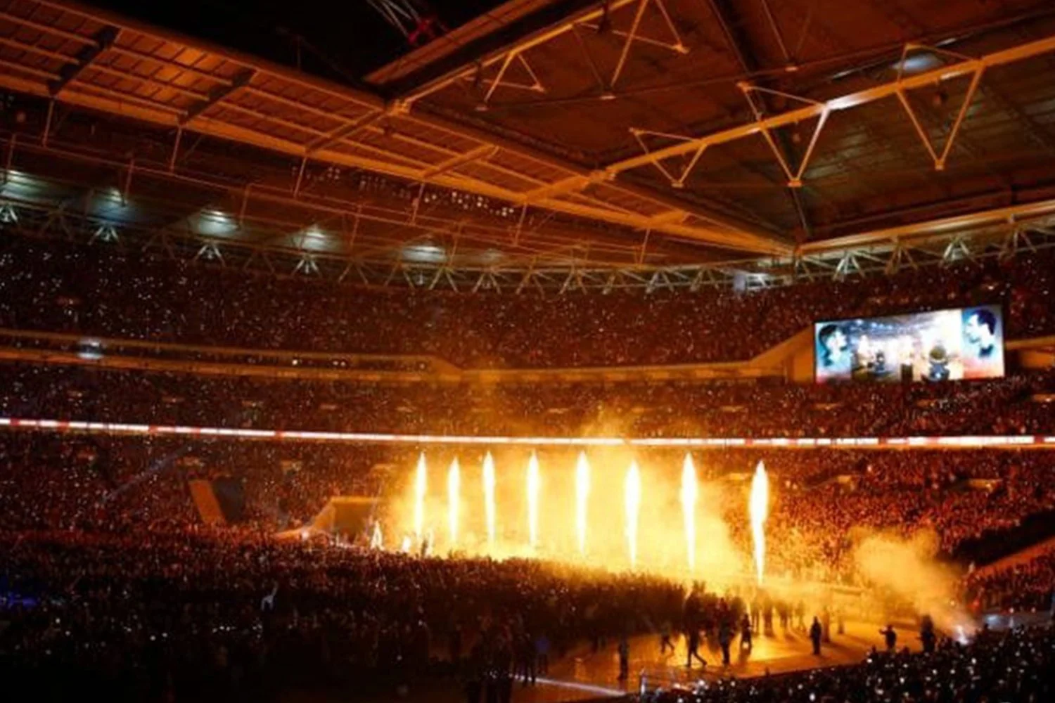 A large indoor arena filled with a crowd watching a fireworks display during a concert or event. Bright orange and yellow fireworks light up the space, with smoke and sparks visible, and a large screen displaying visuals on the right side.