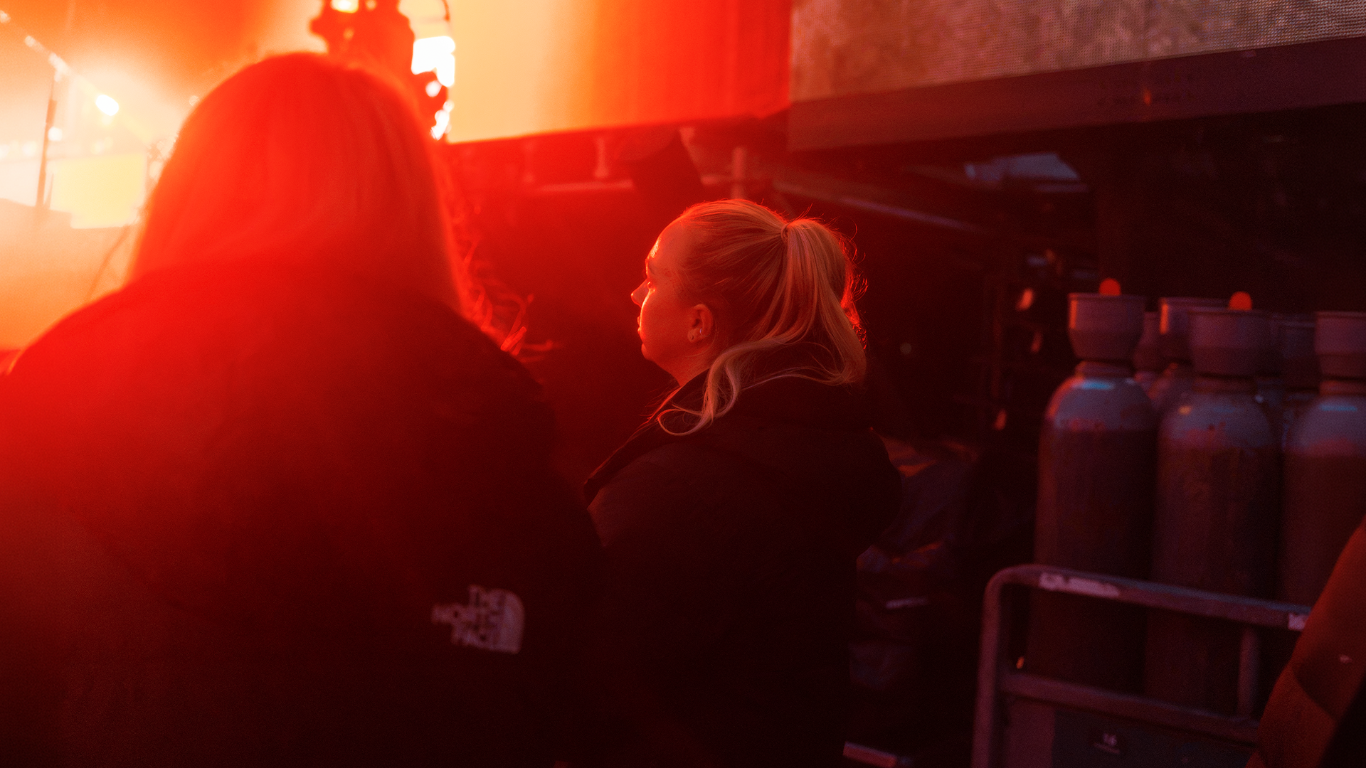 People sitting inside a dark room illuminated by red-orange light, with one woman with blonde hair sitting and looking to the right.