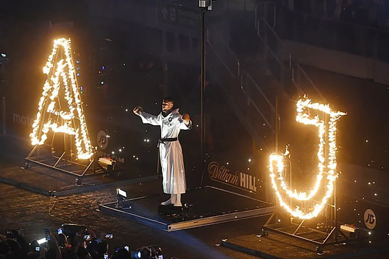 A martial artist in a white uniform performs a routine on a stage with flaming letter formations, a capital 'A' and a lowercase 'u', set against a dark background at William Hill.
