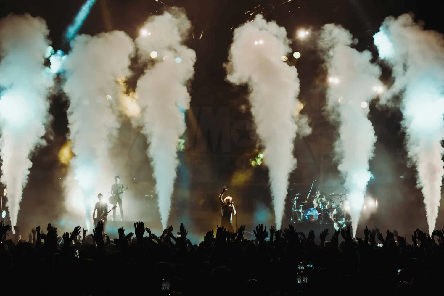 A live concert performance with band members on stage, surrounded by smoke effects and stage lighting, with an audience raising their hands.