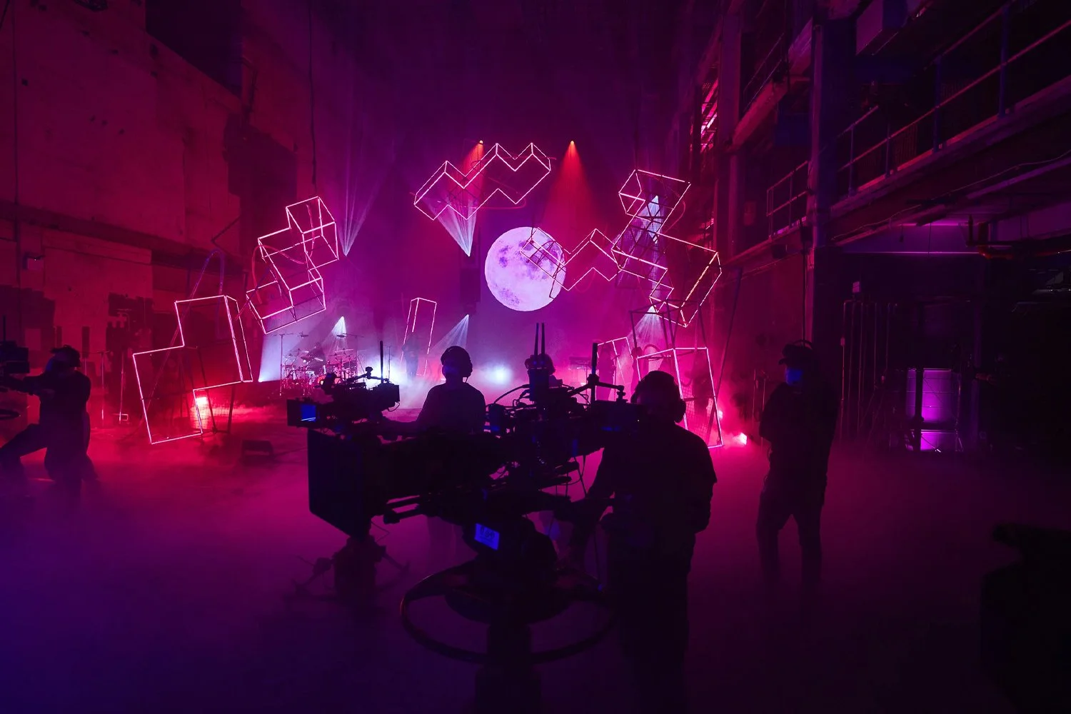 A behind-the-scenes view of a concert or film set with neon geometric light structures, a moon projection, and crew members working in silhouette.