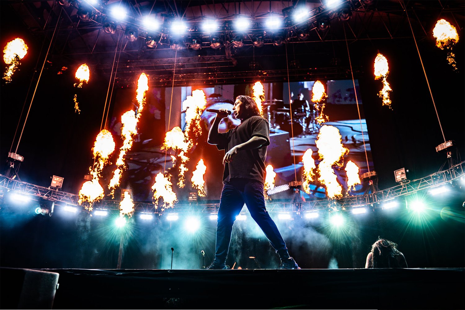 A musician singing into a microphone on stage at a concert with fire effects and bright stage lights.