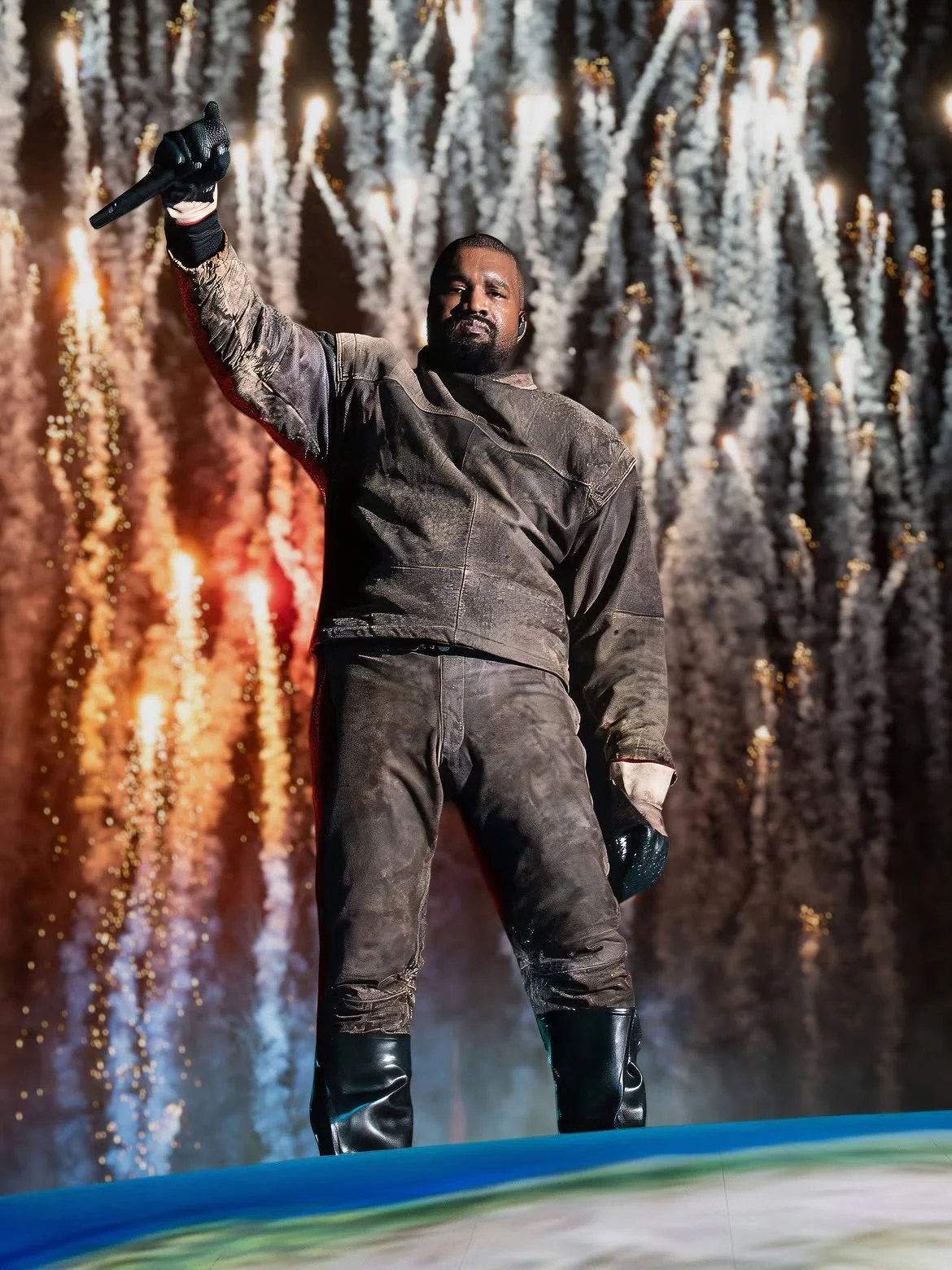 A male performer on stage in front of fireworks displays, wearing a dark, weathered outfit, holding a microphone and a hat, with a confident pose.