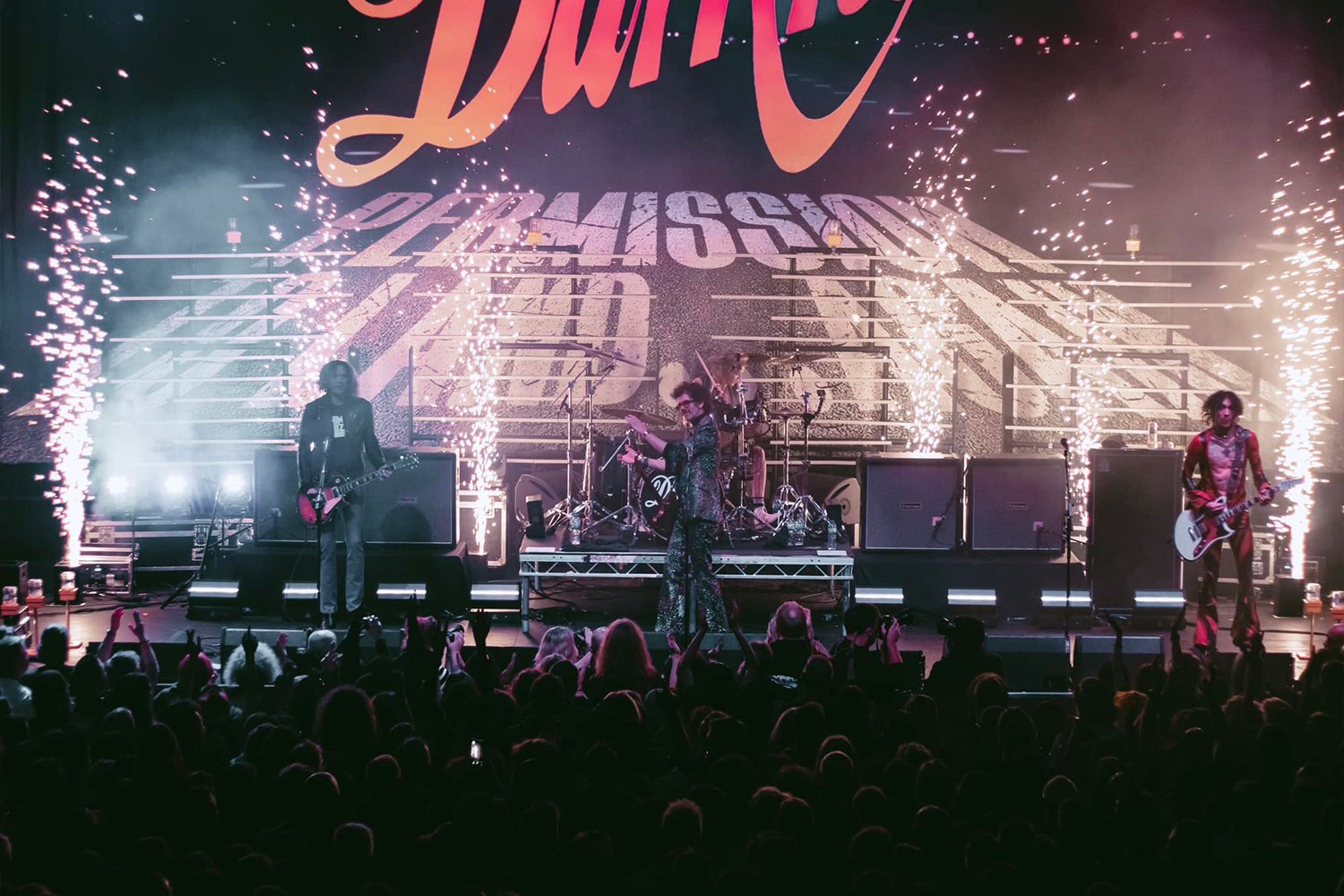 A live band performs on stage with fireworks and bright lights, and an audience is watching in the foreground.