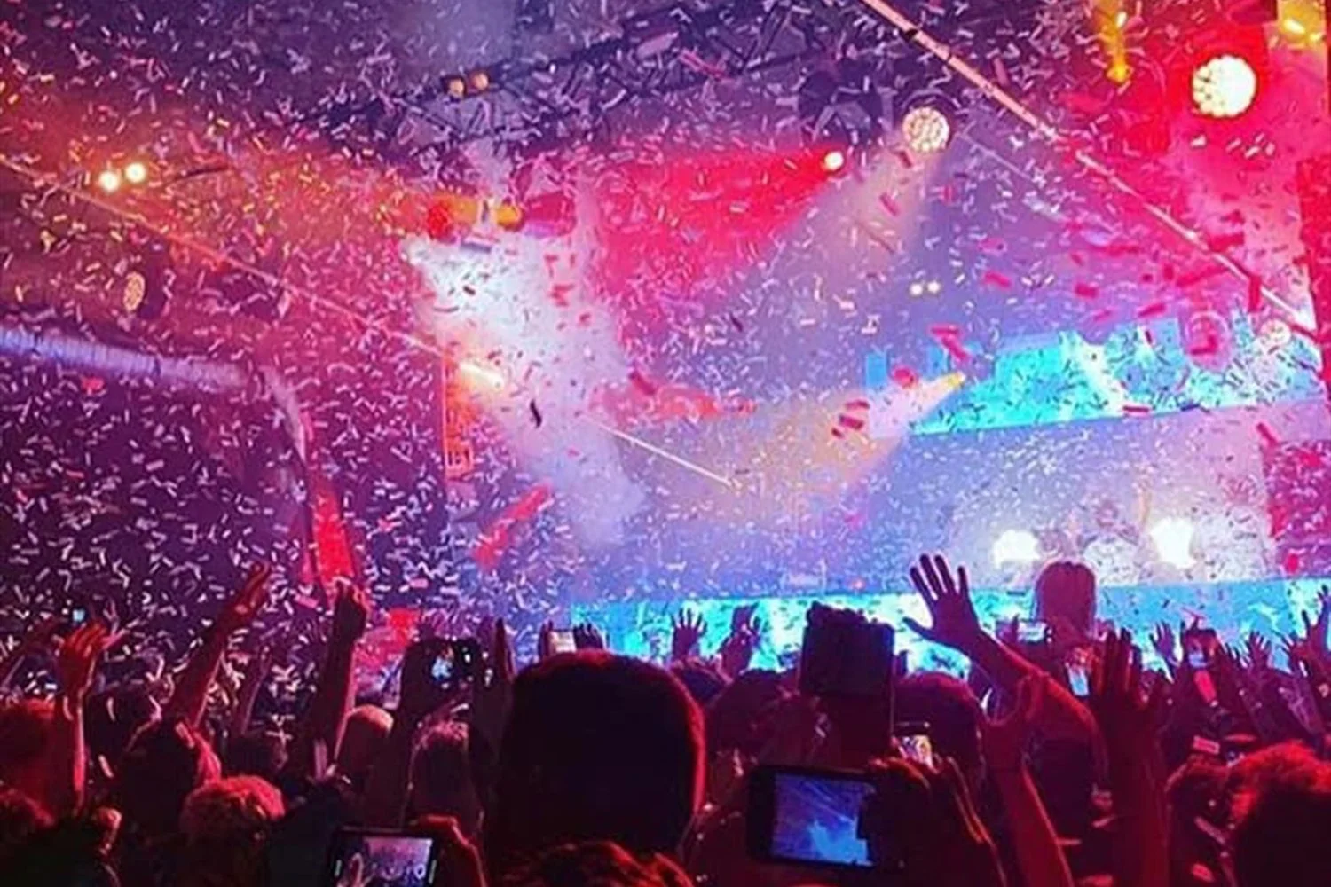 A concert crowd with people raising their hands, confetti falling, colorful stage lights, and a large screen displaying visuals.