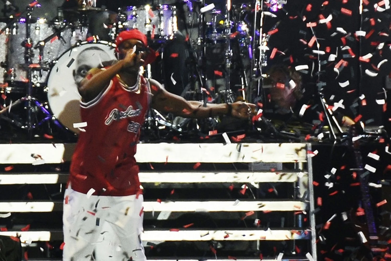 A man in a red basketball jersey and white pants celebrating on stage with red, white, and black confetti falling around him. He is wearing a red cap and sunglasses, with jewelry around his neck, and is making a gesture with his right hand.