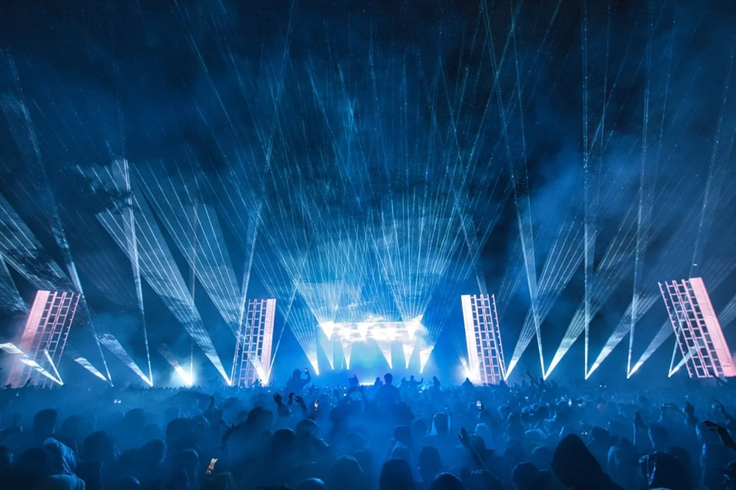 A concert or music event with stage lighting and laser beams, large screen displays, and a crowd of people enjoying the show at night.