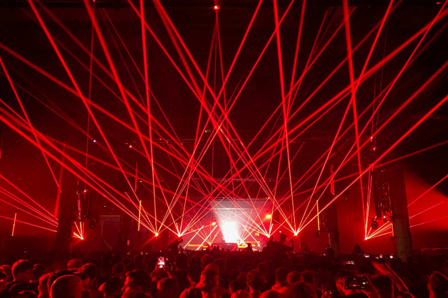 Red laser lights create an intricate pattern above a crowd at a concert or event, with a bright stage and audience in the foreground.