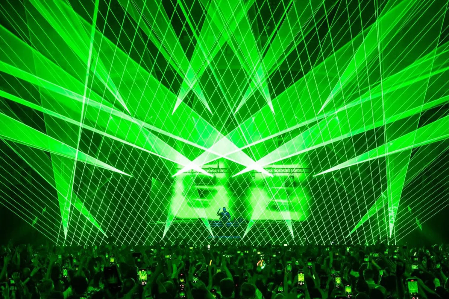 A concert with green laser lights and beams on stage, with a crowd holding up phones to record the performance.