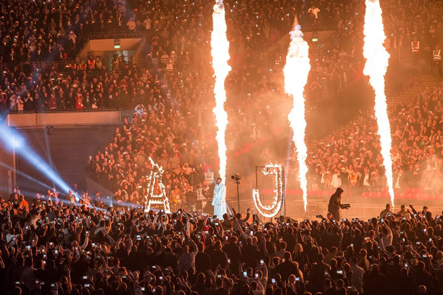 Crowd at a large indoor event with pyrotechnic displays, lasers, and a performer on stage.