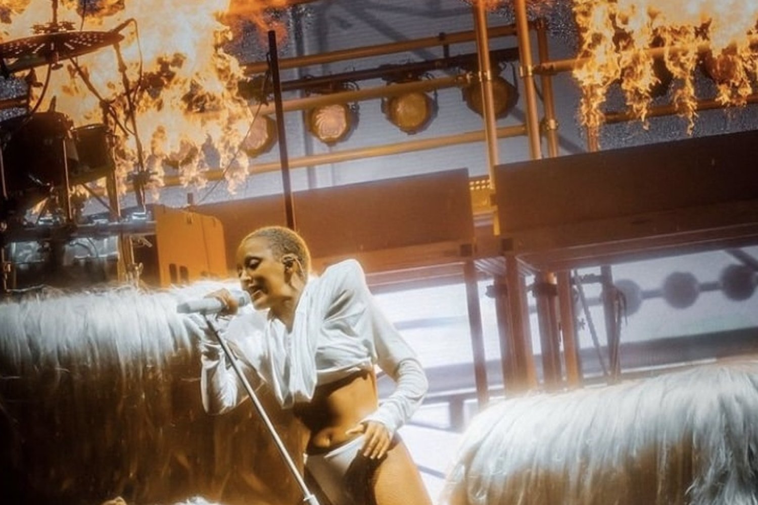 A performer with short hair singing into a microphone during a live concert, with flames and stage lighting in the background.