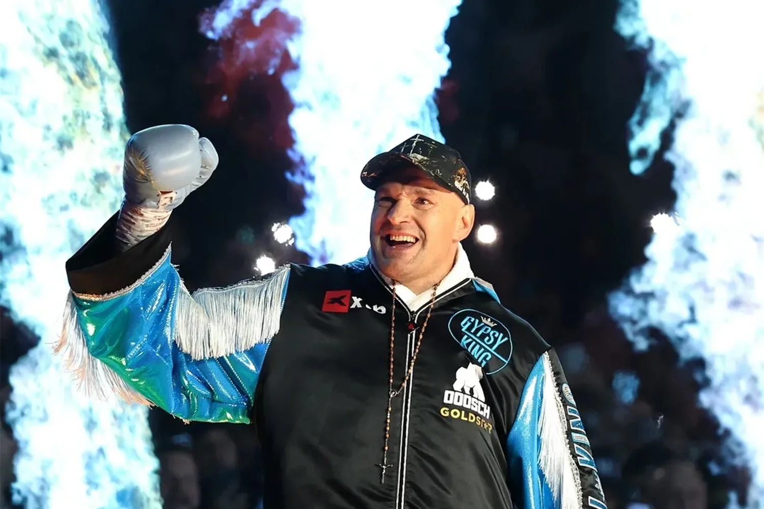 A man wearing a black cap, black jacket with patches, and a colorful shiny sleeve is celebrating with a raised fist in front of a volcanic eruption with bright blue and white ash clouds.
