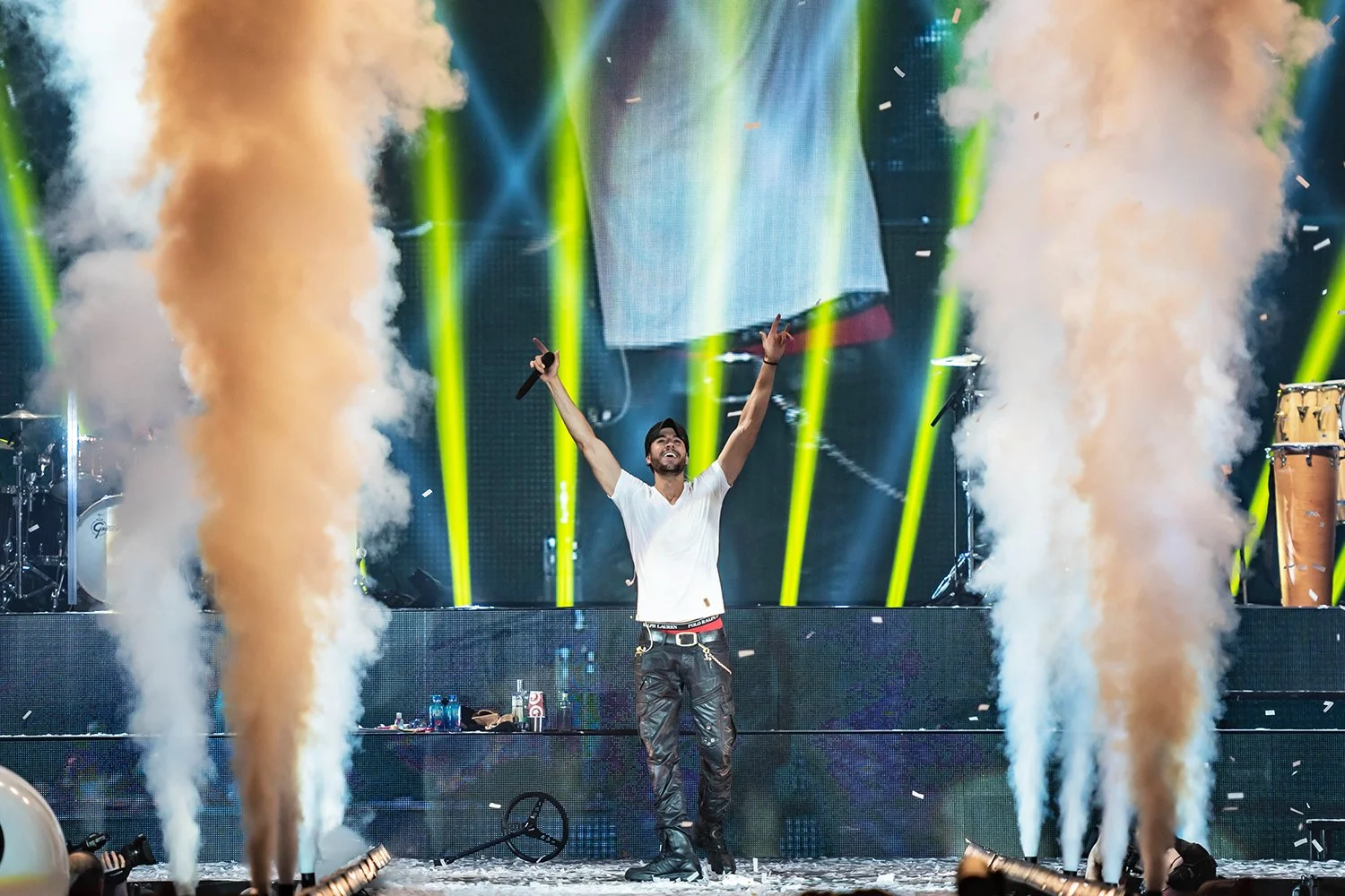 A man on stage with his arms raised, holding a microphone, surrounded by smoke and bright yellow stage lights.