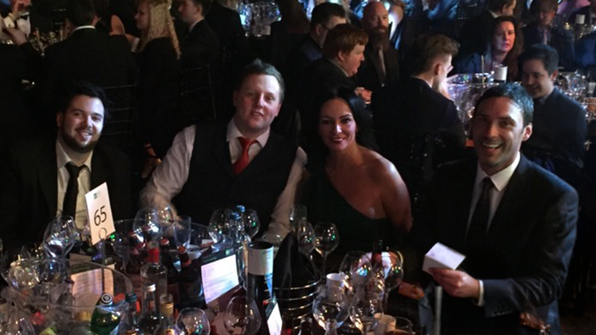 Group of people enjoying a formal dinner at a large banquet table with wine glasses and bottles, in a dimly lit event setting.