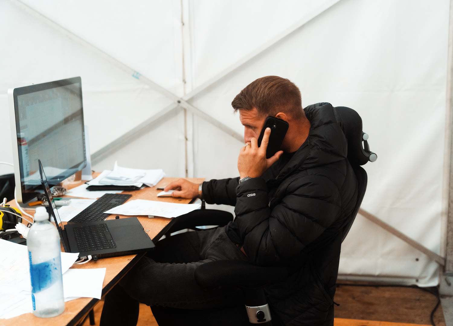 Man sitting at a desk in a white tent, talking on a cellphone, with a desktop computer, keyboard, papers, and a bottle of hand sanitizer on the desk.